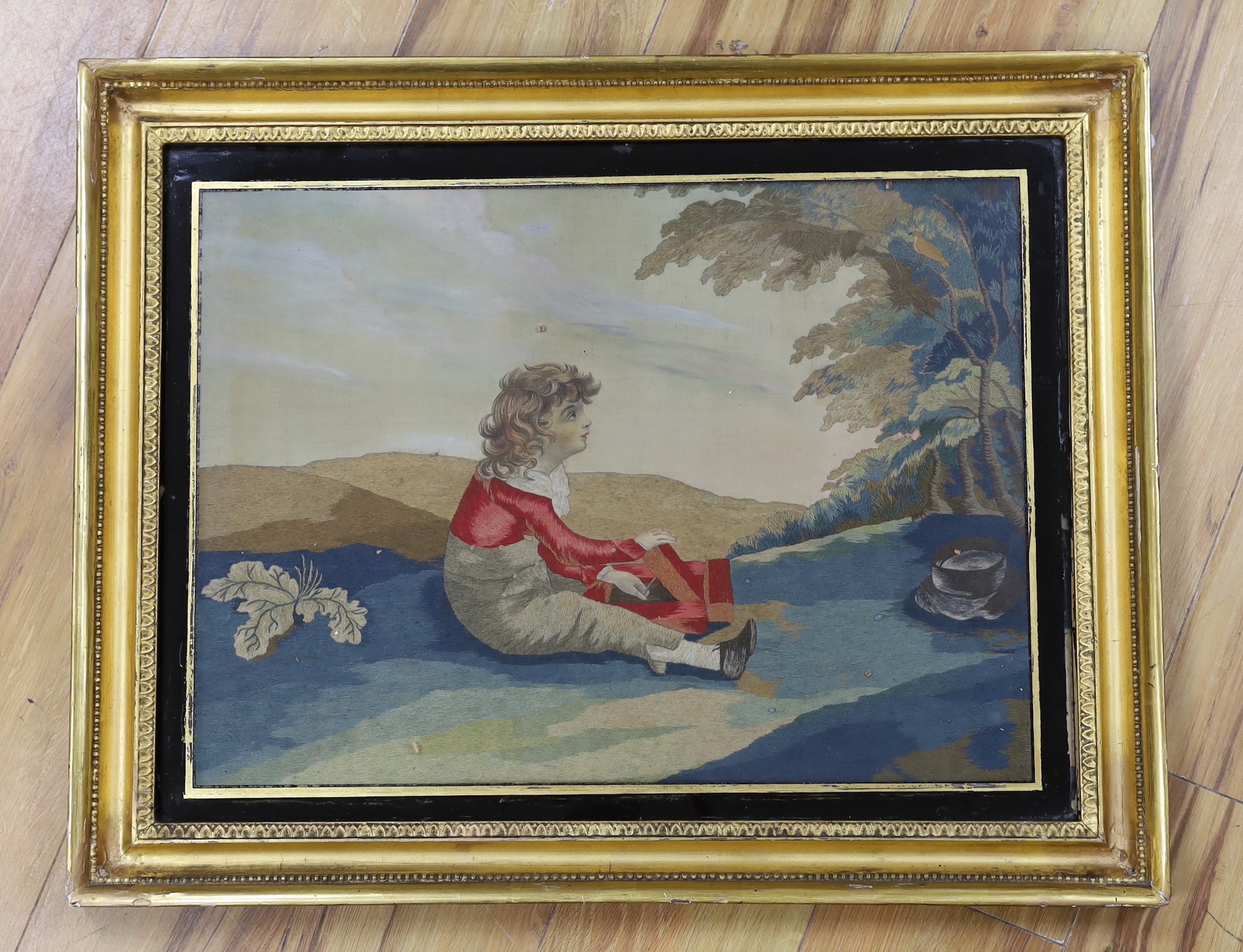 A Regency silk and woolwork picture of a boy sitting under a tree with a bird catcher, 55cm wide x 40cm high